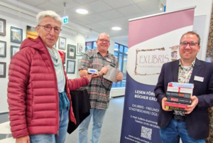 Auf dem Bild sind drei Mitglieder des Fördervereins zu sehen. Eine Person hält eine digitale Waage in der Hand, eine andere Person hält einen Bücherstapel. Im Hintergrund ist das Roll-up des Fördervereins zu sehen.