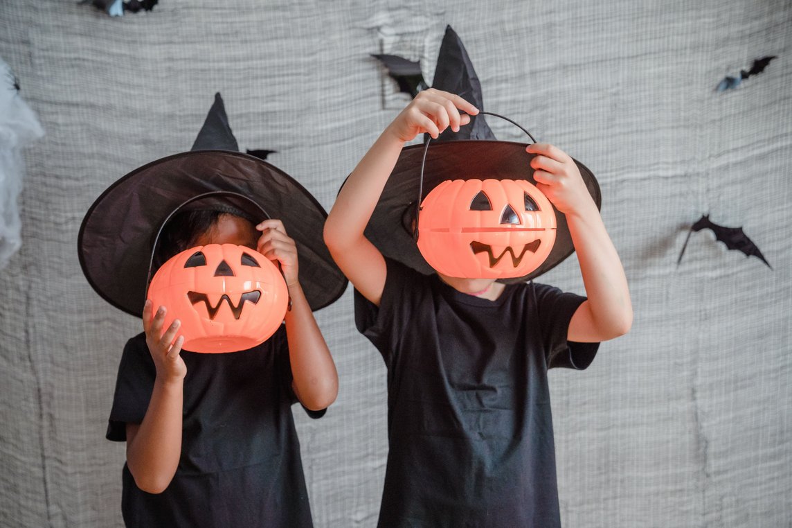 Auf dem Foto halten sich zwei als Hexen verkleidete Kinder jeweils eine Kürbislaterne vor das Gesicht.