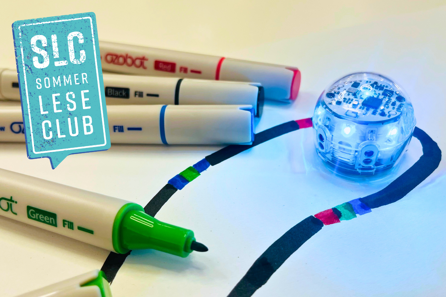 Auf einem weißen Papier ist mit einem schwarzen Marker eine Linie gemalt worden, die von verschiedenen Farbfolgen unterbrochen wird. Auf der Linie steht ein kleiner Ozobot-Roboter, dessen Lichter an sind. Daneben liegen vier verschiedenfarbige Stifte. Einer von ihnen ist geöffnet.