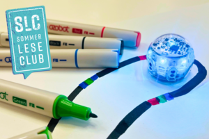 Auf einem weißen Papier ist mit einem schwarzen Marker eine Linie gemalt worden, die von verschiedenen Farbfolgen unterbrochen wird. Auf der Linie steht ein kleiner Ozobot-Roboter, dessen Lichter an sind. Daneben liegen vier verschiedenfarbige Stifte. Einer von ihnen ist geöffnet.