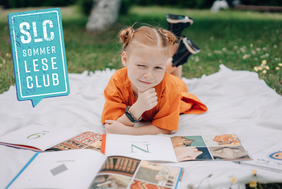 Auf einer Decke liegt ein Mädchen mit roten Haaren und einem orangenen Kleid auf dem Rasen. Sie zwinkert in die Kamera und vor ihr liegen mehrere aufgeschlagene Bücher.